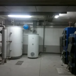 Piwnica w Kłoczewie z zainstalowanymi dwoma białymi zbiornikami na wodę, rurami, niebieskim naczyniem wzbiorczym i niebieskim piecem na paliwo stałe z otwartymi drzwiczkami paleniska.