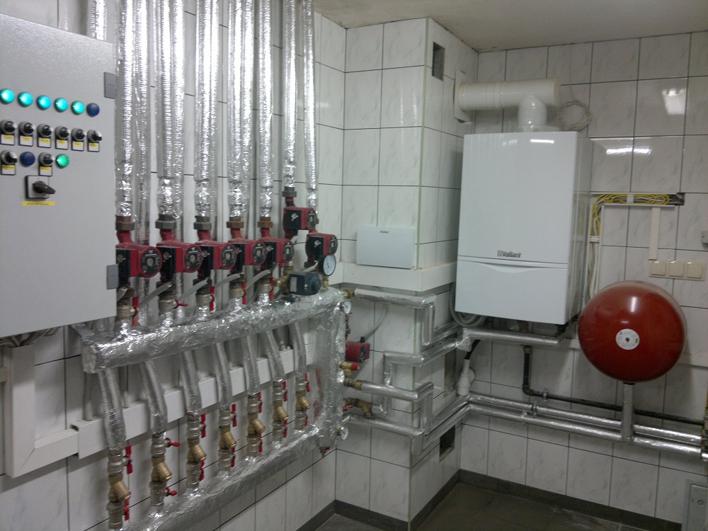 Biały piec gazowy marki Vaillant zamontowany na ścianie z białych płytek, obok czerwony zbiornik wyrównawczy, liczne rury w izolacji termicznej oraz szafa sterownicza z zielonymi diodami.
