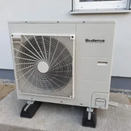 Zewnętrzna jednostka pompy ciepła marki Buderus, zamontowana na betonowej podstawie z użyciem podkładek antywibracyjnych, widoczna ściana budynku z oknem.