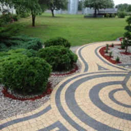 Zakrzywiony chodnik z kostki brukowej w odcieniach beżu i grafitu, obrzeża z kamyków i zieleni. Widok na trawnik i drzewa w tle, drzwi wejściowe po prawej.