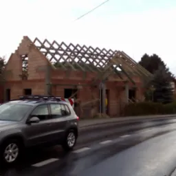 Wznoszenie domu murowanego z czerwonej cegły, widoczna konstrukcja dachu z drewnianych belek, na pierwszym planie szary samochód osobowy na mokrej ulicy.