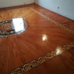 Lakierowana drewniana podłoga z intarsją w kształcie rozety i bordiury z motywem roślinnym, widoczna z perspektywy narożnika pokoju.