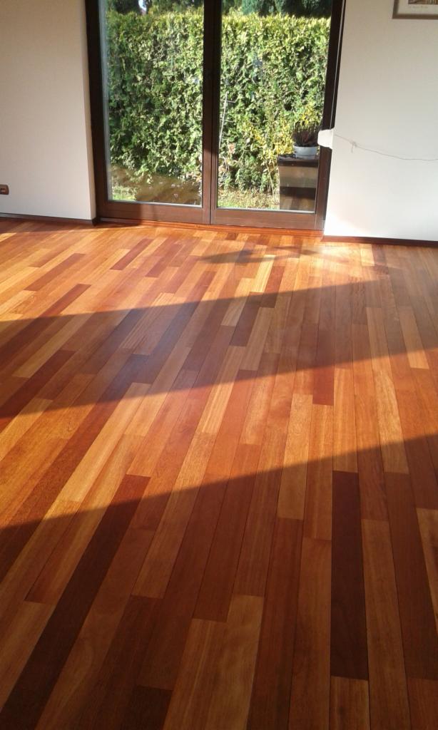 Ułożona podłoga z egzotycznego drewna, widoczne naturalne usłojenie i ciepłe odcienie brązu, duże okno tarasowe z widokiem na zielony żywopłot w tle.