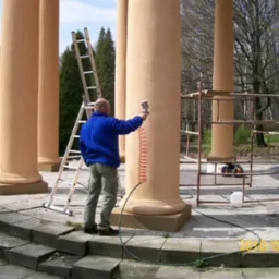 Pracownik w niebieskiej bluzie nanosi tynk maszynowy na kolumnę w altanie za pomocą pistoletu natryskowego. Widoczna drabina i rusztowanie.