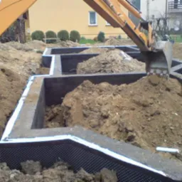 Fundamenty domu w trakcie budowy, zabezpieczone izolacją, z wykopaną ziemią. Koparka częściowo widoczna w tle. Prace ziemne w toku.