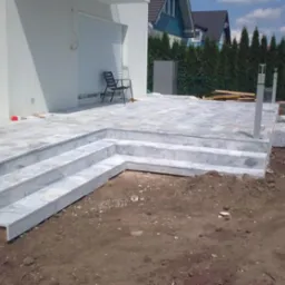 Wykończony taras z marmurowymi schodami, z widocznym fragmentem domu i zieleni w tle. Widoczne lekkie niedoróbki, ziemia wokół.