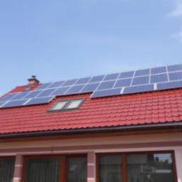 Czerwony dach z panelami słonecznymi i oknem dachowym na tle błękitnego nieba. Widok na dom z panelami fotowoltaicznymi.