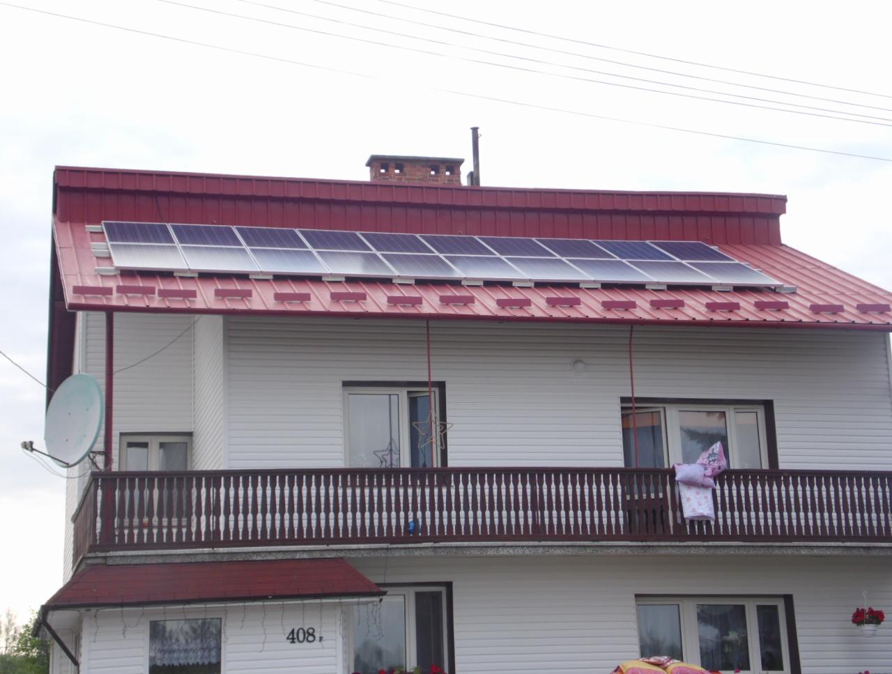 Panele słoneczne na czerwonym dachu dwupiętrowego domu z białą elewacją. Widoczna antena satelitarna i dekoracje na balkonie.