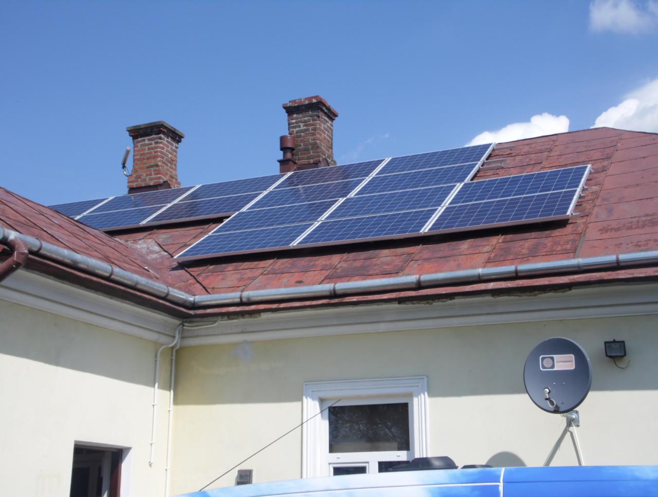 Instalacja paneli słonecznych na dachu domu z ceglaną dachówką i dwoma kominami. Widoczna antena satelitarna na ścianie budynku.