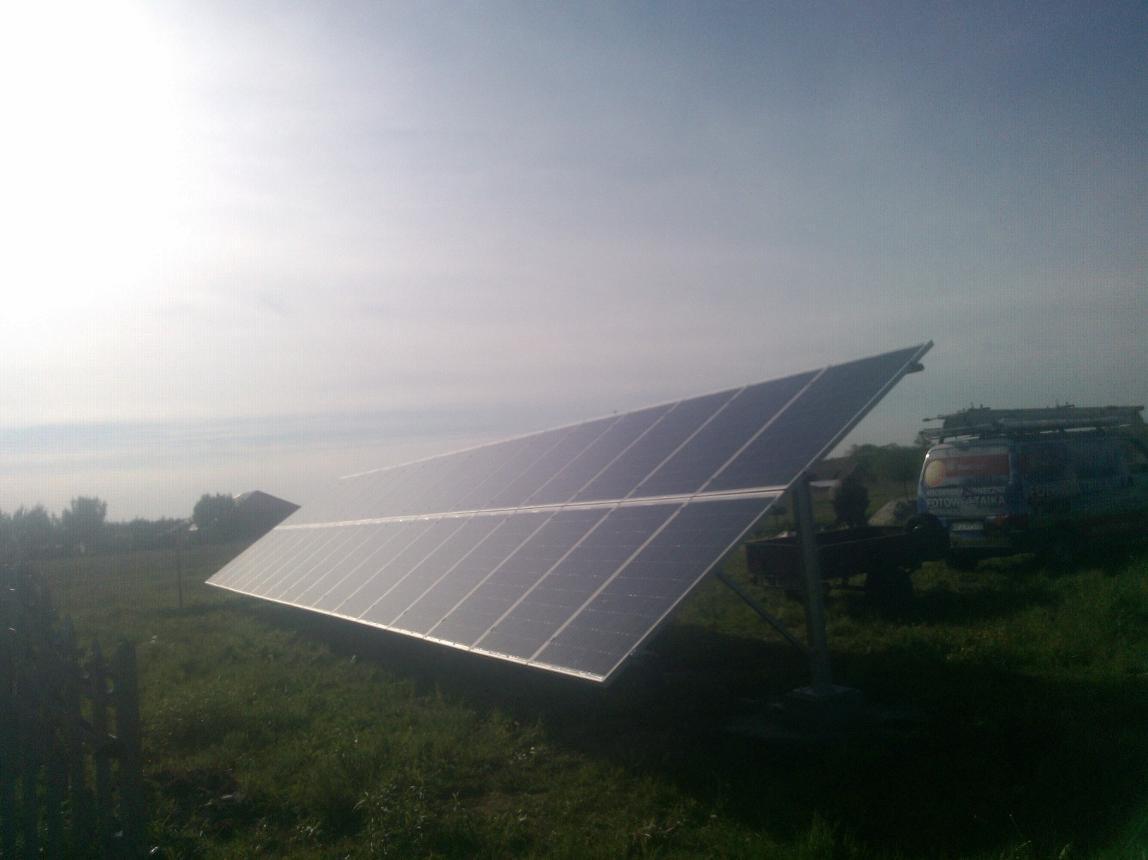 Panel fotowoltaiczny na łące, widok z dołu. Błękitne niebo w tle, obok zaparkowany bus z napisem. Instalacja solarna na trawie, w oddali zabudowania.