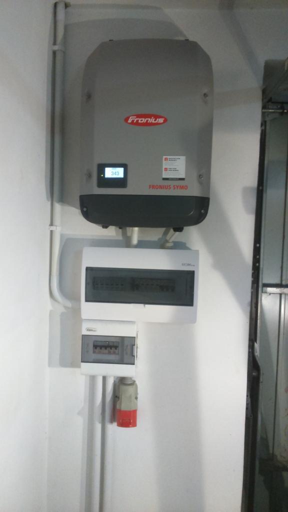 Szary falownik Fronius Symo zamontowany na białej ścianie, pod nim dwie rozdzielnice elektryczne i gniazdo siłowe. Na wyświetlaczu falownika widoczna wartość 343.
