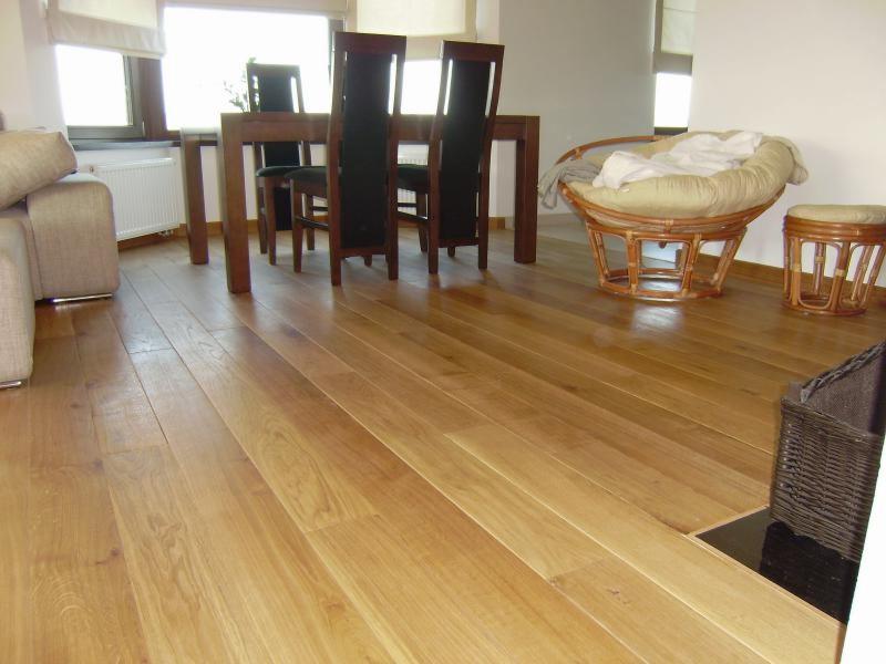 Jasna, drewniana podłoga z wyraźnym usłojeniem w salonie. W tle stół z krzesłami, wiklinowy fotel i pufa. Naturalne światło wpada przez okno.