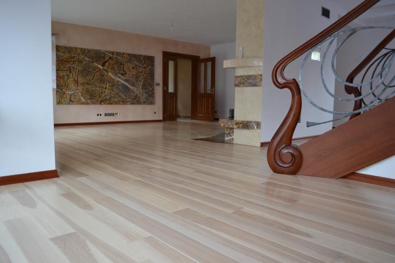 Jasna, drewniana podłoga z wyraźnym usłojeniem, widok z niskiej perspektywy. W tle drzwi dwuskrzydłowe, fragment schodów z ozdobną balustradą i dekoracyjny panel na ścianie.