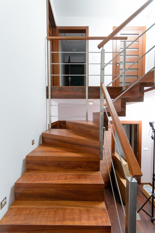 Drewniane schody spiralne z metalową balustradą, widoczne drzwi na dwóch poziomach. Ciepła kolorystyka drewna kontrastuje z bielą ścian. Oświetlenie punktowe na schodach.