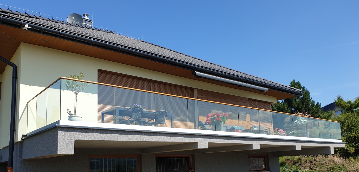 Szklana balustrada z drewnianą poręczą na balkonie nowoczesnego domu w Dzięgielowie, widok z dołu, na balkonie stolik i krzesła.