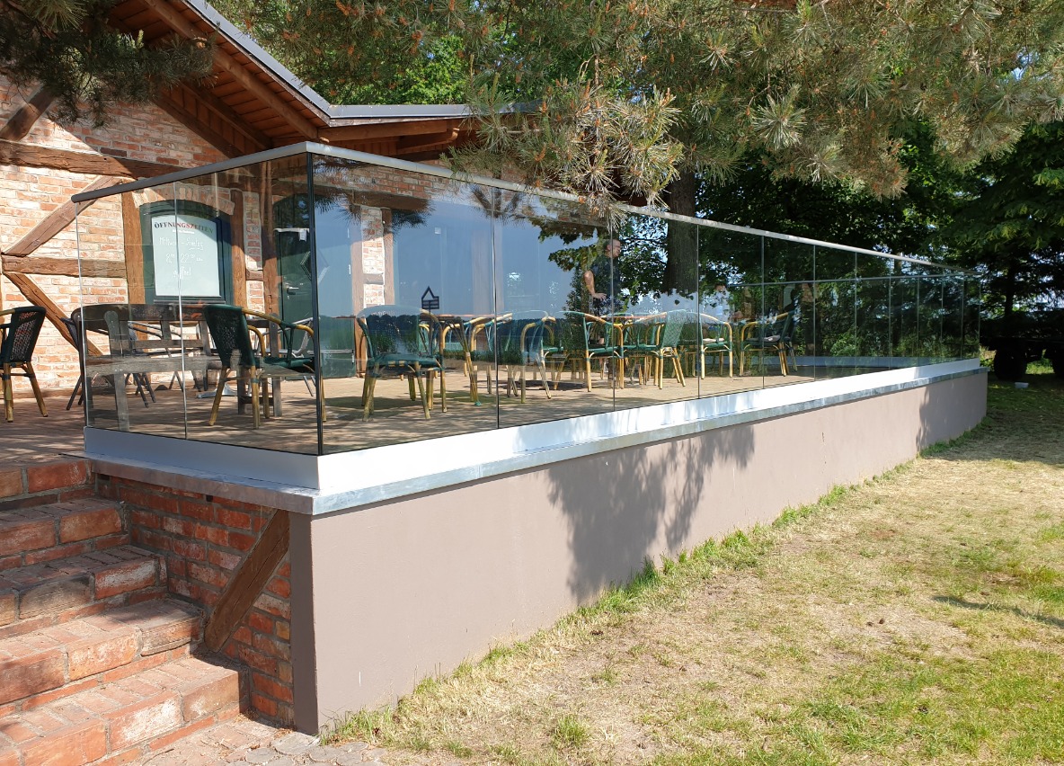 Szklana barierka na tarasie restauracji w stylu rustykalnym z widocznymi ceglanymi schodami i drewnianą konstrukcją budynku w tle.