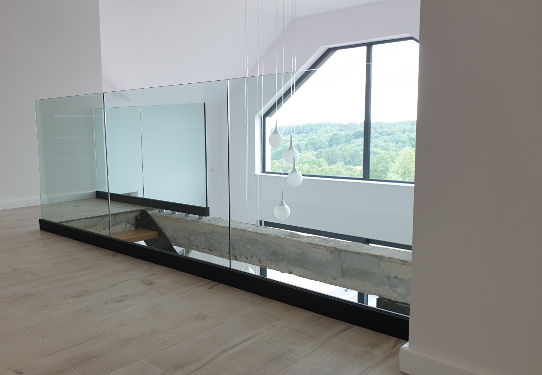 Nowoczesna balustrada ze szkła na antresoli, z widocznym betonowym elementem konstrukcyjnym i oknem z widokiem na zalesione wzgórza.