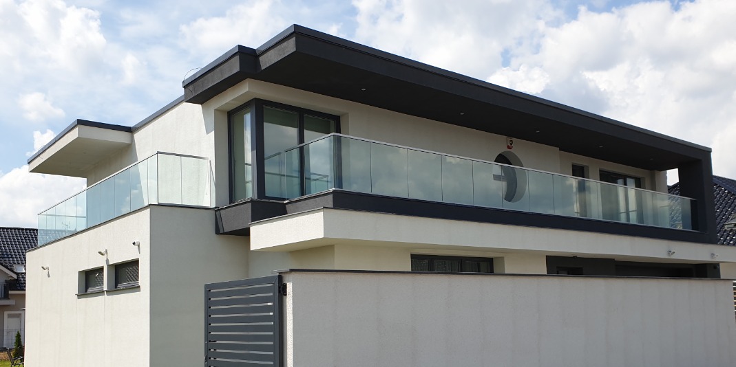 Nowoczesny dom z białymi ścianami i czarnymi akcentami, z balkonem zabezpieczonym szkłem, widok z poziomu ulicy.