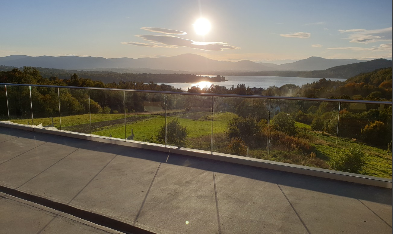 Szklana balustrada balkonowa z widokiem na jezioro i góry w Żywcu w promieniach zachodzącego słońca.