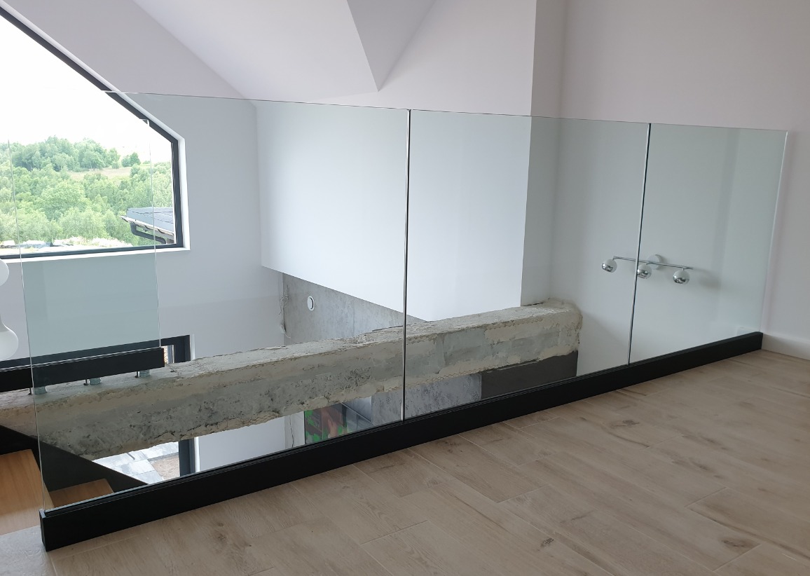 Szklana bariera ochronna na antresoli z widokiem na las, czarna rama, drewniana podłoga, surowy betonowy element konstrukcyjny.