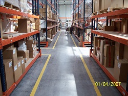 Szary betonowy korytarz w magazynie z żółtymi liniami wyznaczającymi ścieżki, regały po obu stronach wypełnione kartonami.