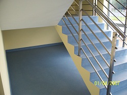 Klatka schodowa z niebieskimi płytkami na schodach i szarą, jednolitą powierzchnią podłogi. Metalowa barierka z poziomymi prętami. Ściany pomalowane na jasnożółty kolor. Widoczny fragment daty...