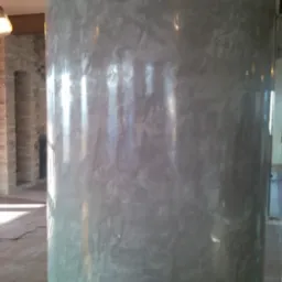 Okrągła kolumna z szarym tynkiem dekoracyjnym imitującym beton, widoczny fragment kamiennej ściany w tle.