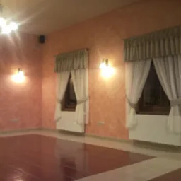 Pokój z pomarańczowymi ścianami o nieregularnej fakturze, trzema oknami z białymi firankami i brązowymi zasłonami, brązową podłogą z jasnymi obramowaniami, oświetlony przez żyrandol i kinkiety.