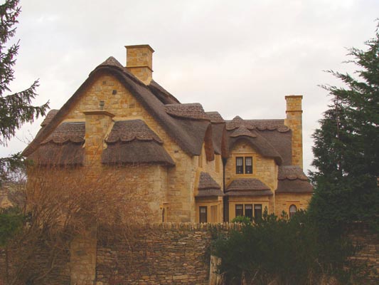 Kamienny dom z dachem krytym strzechą i kominami, otoczony zielenią i niskim kamiennym murem. Dach ma dekoracyjne wykończenie krawędzi.