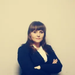 Portret kobiety w ciemnej marynarce i białej koszuli, z założonymi rękami na tle jasnej ściany.