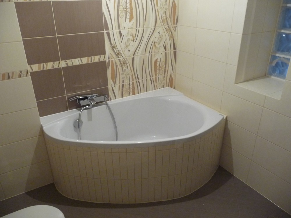 Narożna wanna obudowana beżowymi płytkami w łazience z mozaiką i wzorzystymi płytkami na ścianach, bateria chromowana z wężem.