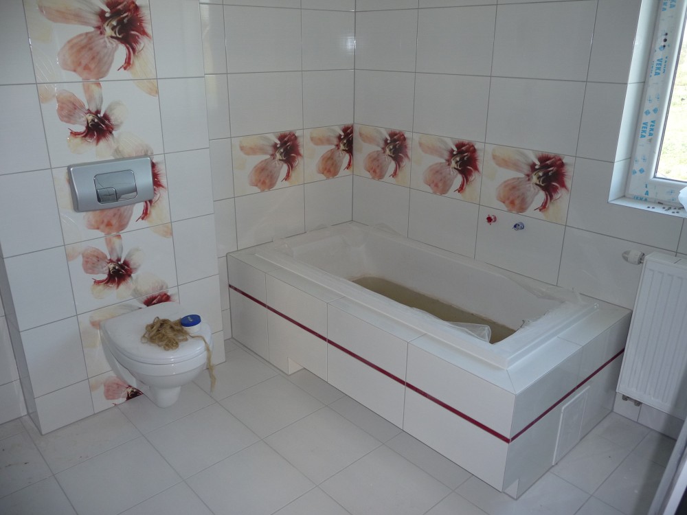 Wykończona łazienka z białymi płytkami zdobionymi motywem kwiatowym, zabudowana wanna z zabezpieczającą folią, podwieszana toaleta z resztkami włosów i fragment grzejnika.