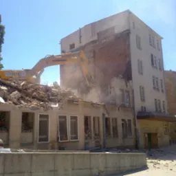 Żółta koparka Liebherr na gruzach budynku podczas rozbiórki, z widocznym fragmentem sąsiedniej kamienicy i drzewem w tle, w słoneczny dzień.