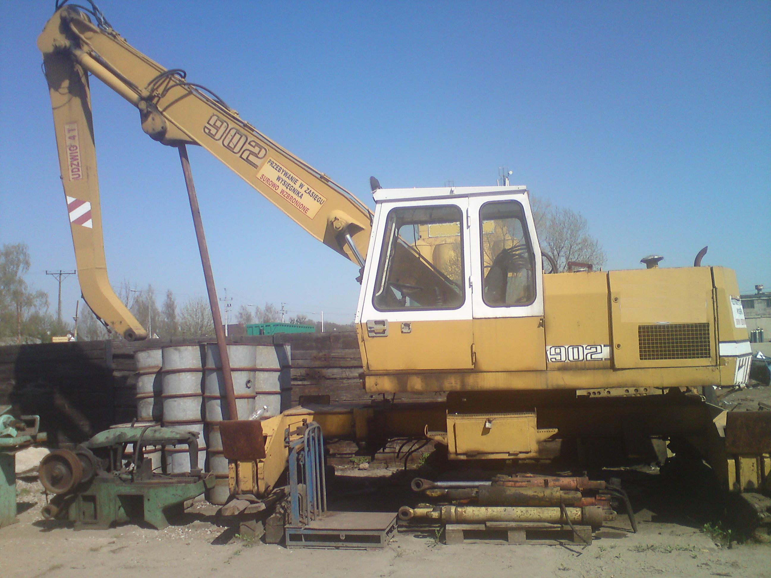 Żółta ładowarka hydrauliczna Liebherr 902, z widocznym wysięgnikiem z napisem ostrzegawczym, stoi na placu budowy obok sterty metalowych beczek i innych maszyn, w tle błękitne niebo.