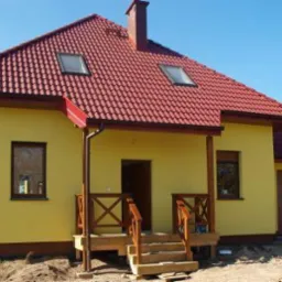 Żółty dom z czerwonym dachem w słoneczny dzień. Widoczny fragment tarasu i garaż. Dom jednorodzinny z poddaszem użytkowym i oknami dachowymi.