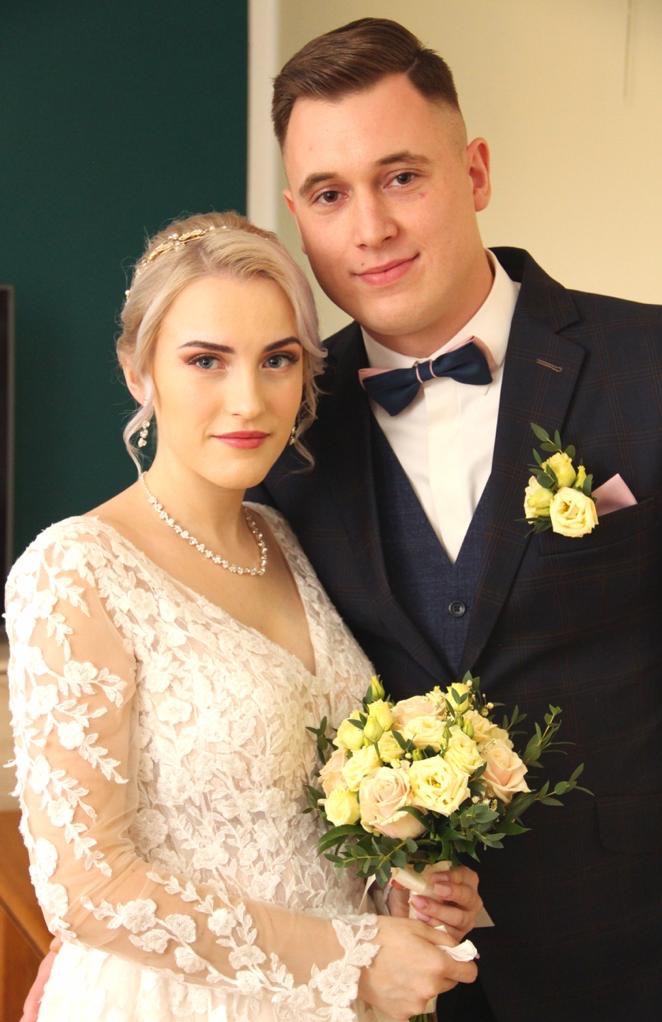 Para młoda w zbliżeniu: panna młoda w koronkowej sukni z bukietem, pan młody w garniturze z muszką i butonierką. Klasyczna elegancja, stonowane kolory, ciepłe oświetlenie.