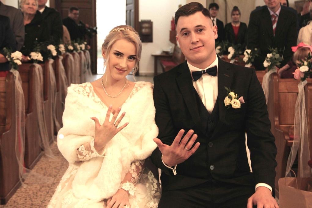 Uśmiechnięta para młoda prezentuje obrączki w kościele w Sopocie. Panna młoda w sukni i futerku, pan młody w czarnym garniturze z muchą. Dekoracje kwiatowe na ławkach.