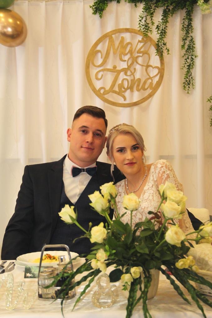 Uśmiechnięta para młoda siedzi przy stole z bukietem białych róż. W tle dekoracja 'Mąż i Żona' na białym tle. Pan w czarnym garniturze, pani w koronkowej sukni.