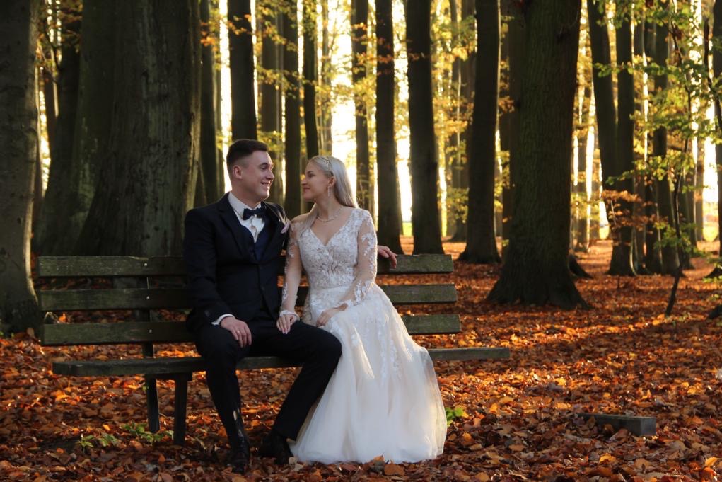 Para młoda siedzi na ławce w lesie jesienią, w tle wysokie drzewa. Pan młody w eleganckim garniturze, panna młoda w białej sukni z koronką. Romantyczna sesja ślubna.