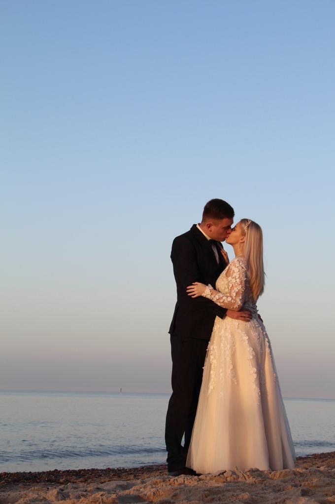 Para młoda w czarnym garniturze i sukni ślubnej z koronkowymi aplikacjami, całująca się na piaszczystej plaży o zachodzie słońca. Romantyczny moment.