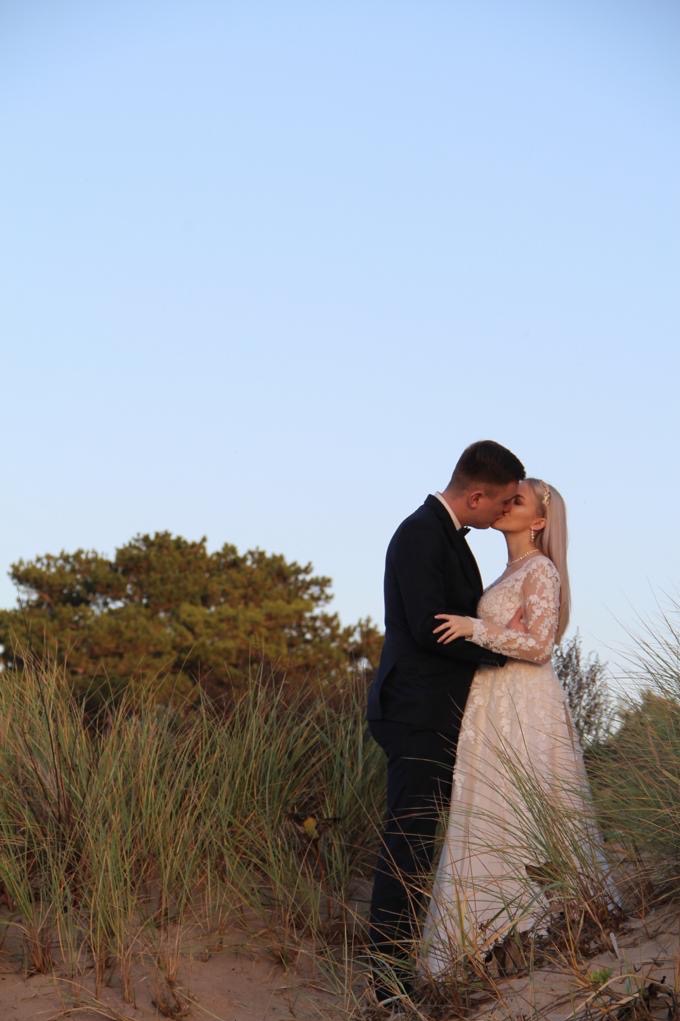 Para młoda w objęciach, całująca się na tle piasku i traw, w delikatnym oświetleniu. Suknia z koronkowymi rękawami. Ujęcie z dołu podkreśla intymność chwili.