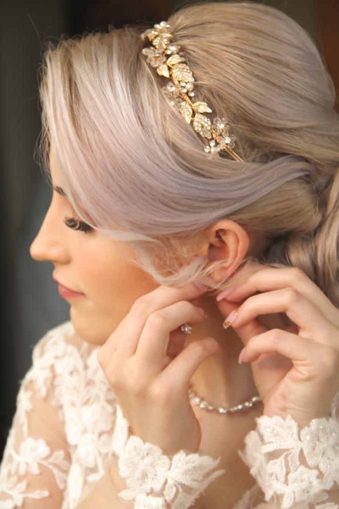 Panna młoda z platynowymi włosami w złotej ozdobie i koronkowej sukni zapina kolczyk. Profil, delikatny makijaż, perłowy naszyjnik, manicure.