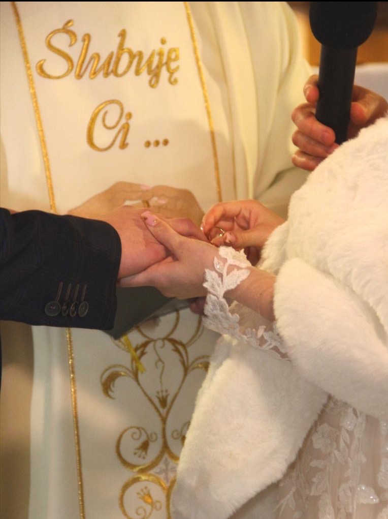 Moment zakładania obrączki podczas ceremonii ślubnej, widoczne dłonie pary młodej i fragment stroju księdza. Na pierwszym planie rękawiczki panny młodej.