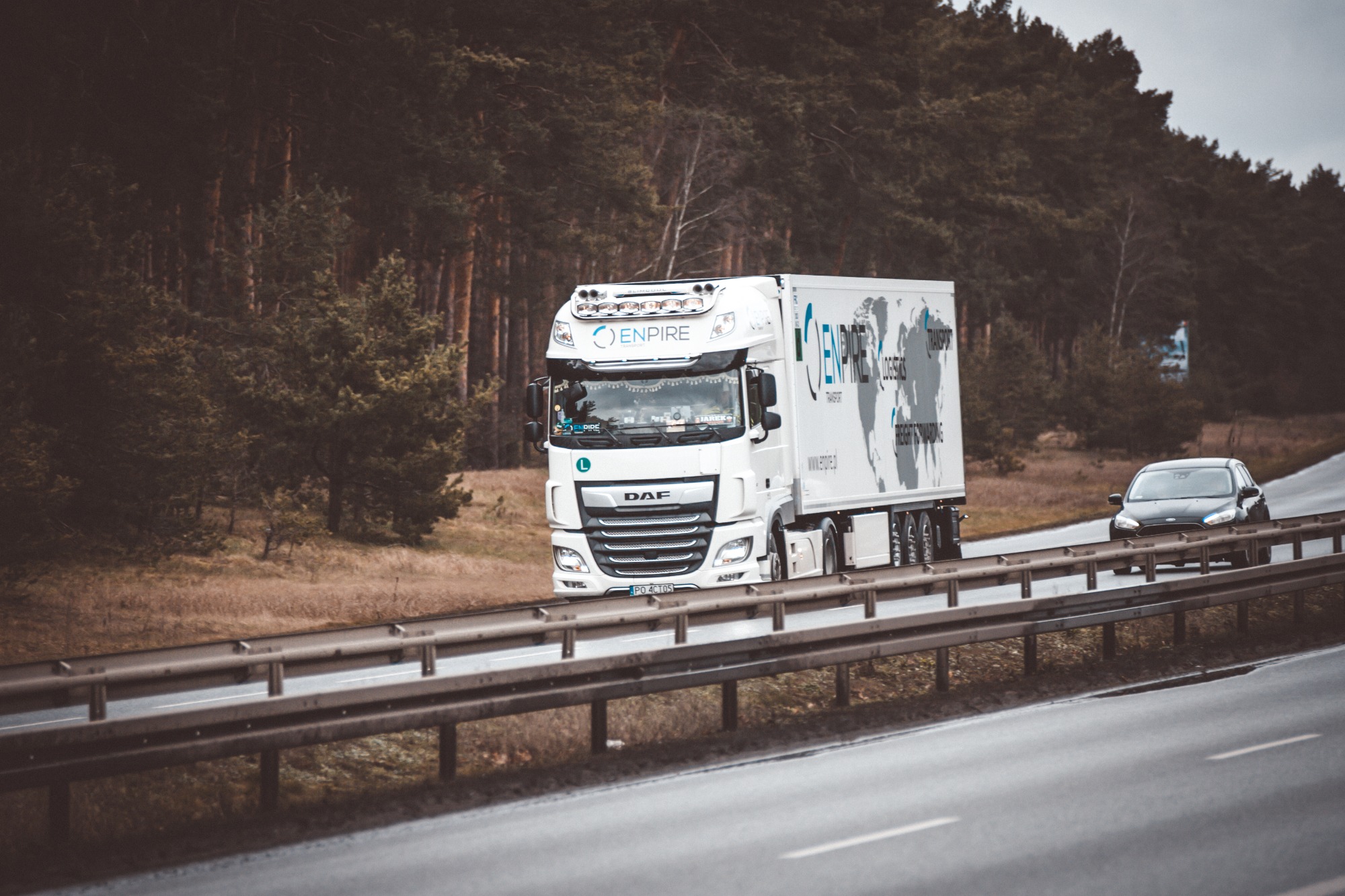 Biała ciężarówka DAF z naczepą, oklejona grafiką mapy świata i logo firmy Enpire Logistics, jadąca autostradą na tle lasu, z widocznym samochodem osobowym.