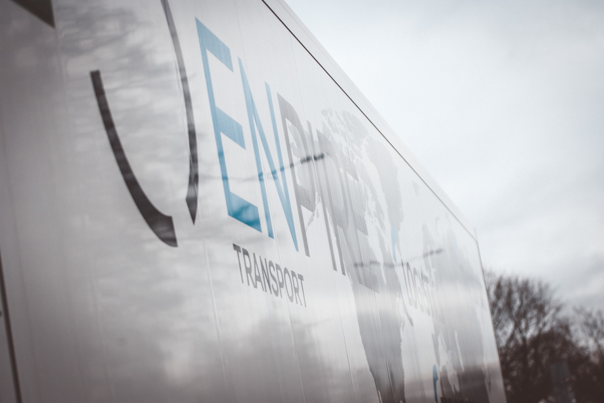 Biała naczepa z niebiesko-czarnym logo firmy transportowej i mapą świata w tle, widziana pod kątem z odbiciem nieba.