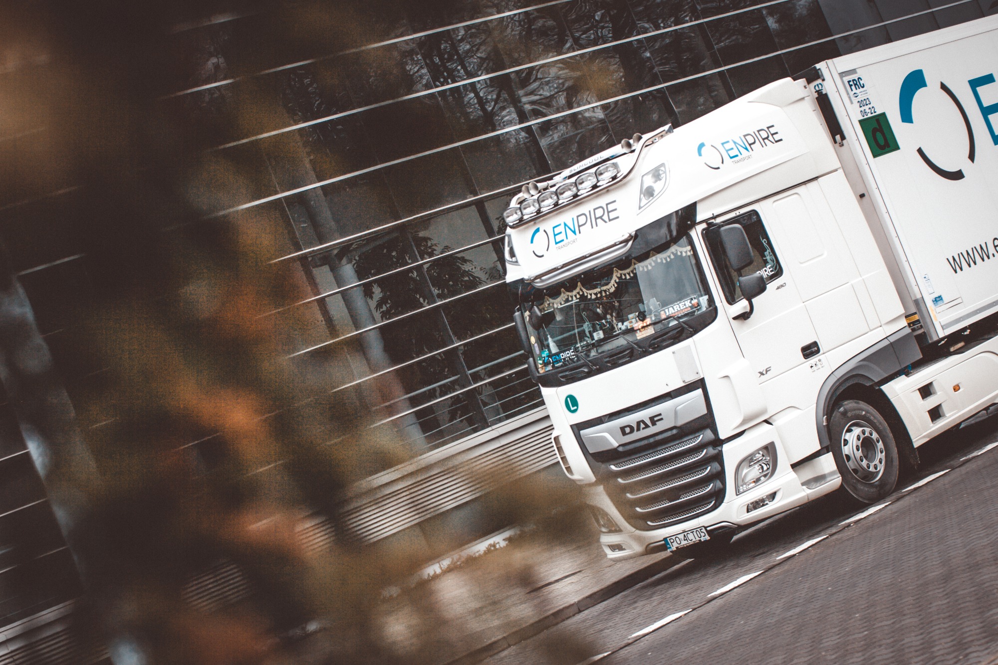 Biała ciężarówka marki DAF z logo firmy Enpire Transport na tle nowoczesnego budynku z przeszkloną fasadą, widok pod kątem z perspektywy żabiej, dekoracje w kabinie kierowcy.