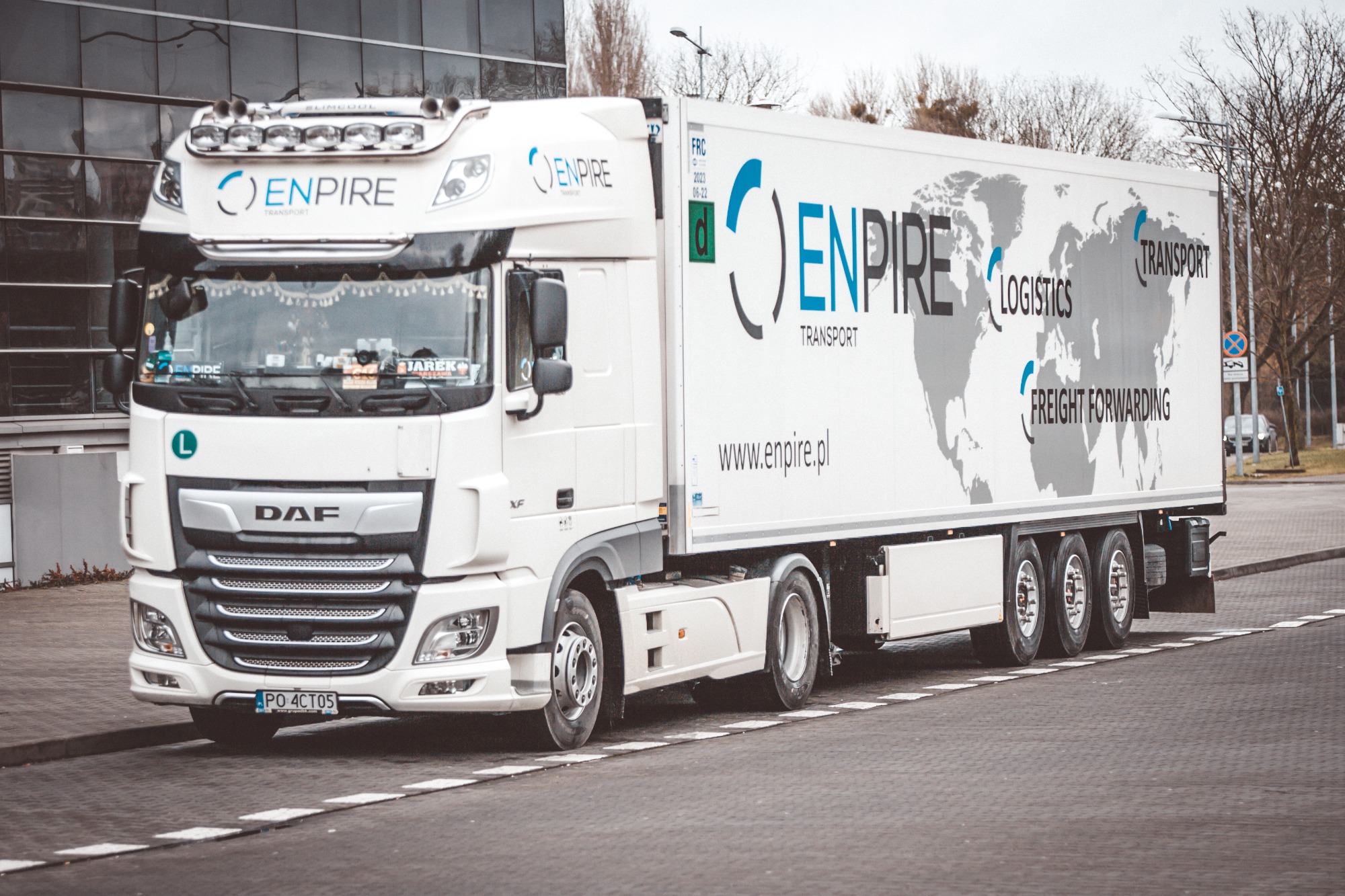 Biała ciężarówka marki DAF z naczepą, na której widnieje mapa świata i logo firmy Enpire Transport, zaparkowana na parkingu przed nowoczesnym budynkiem z przeszkloną fasadą.