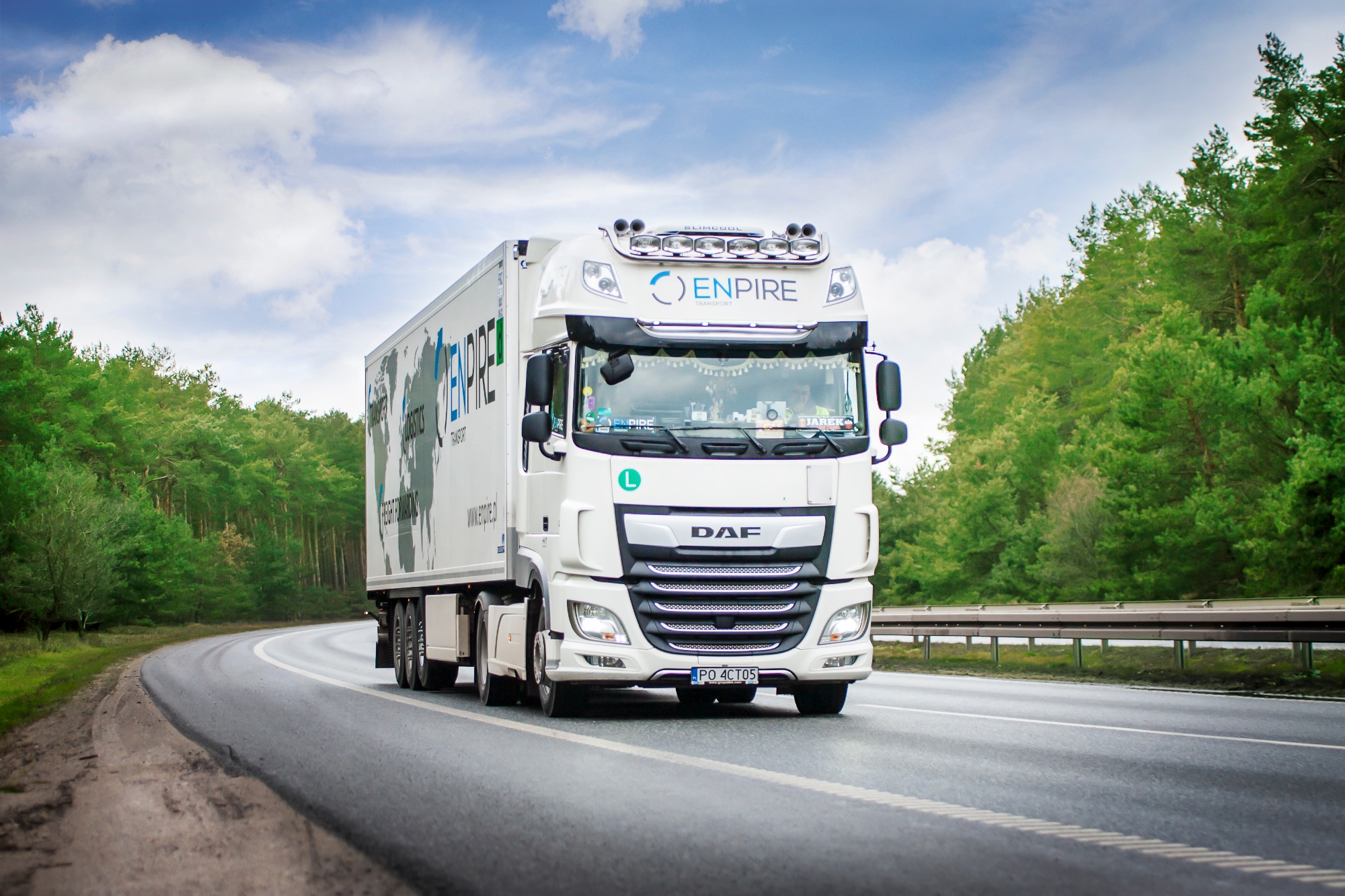 Biała ciężarówka DAF z naczepą firmy Enpire Logistics na asfaltowej drodze wijącej się przez las, błękitne niebo z chmurami w tle.