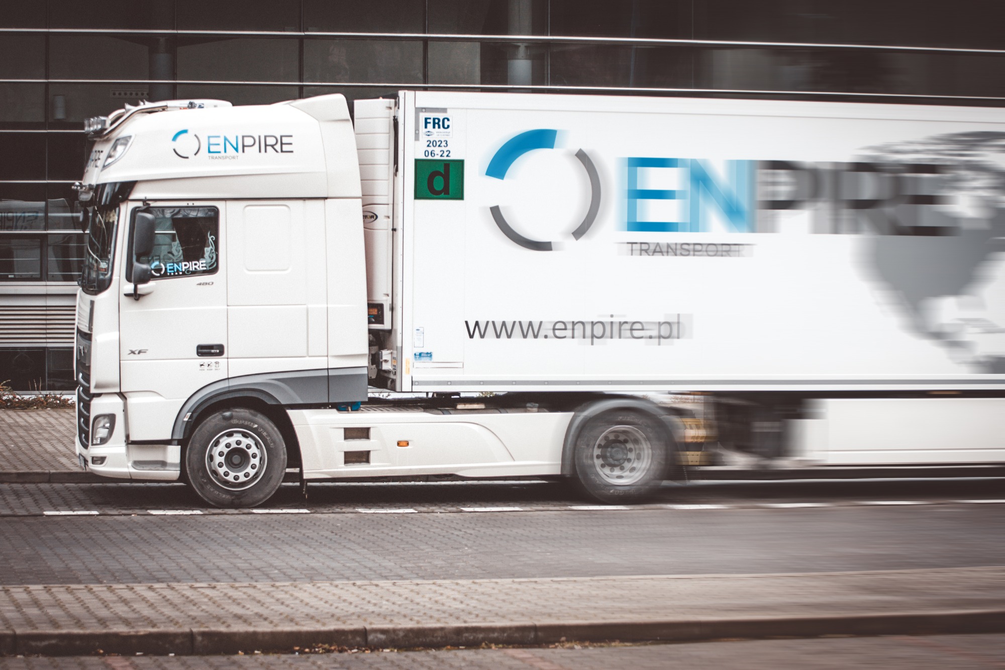 Biała ciężarówka z logo Enpire Transport w ruchu, widoczny adres strony internetowej na naczepie, budynek w tle.
