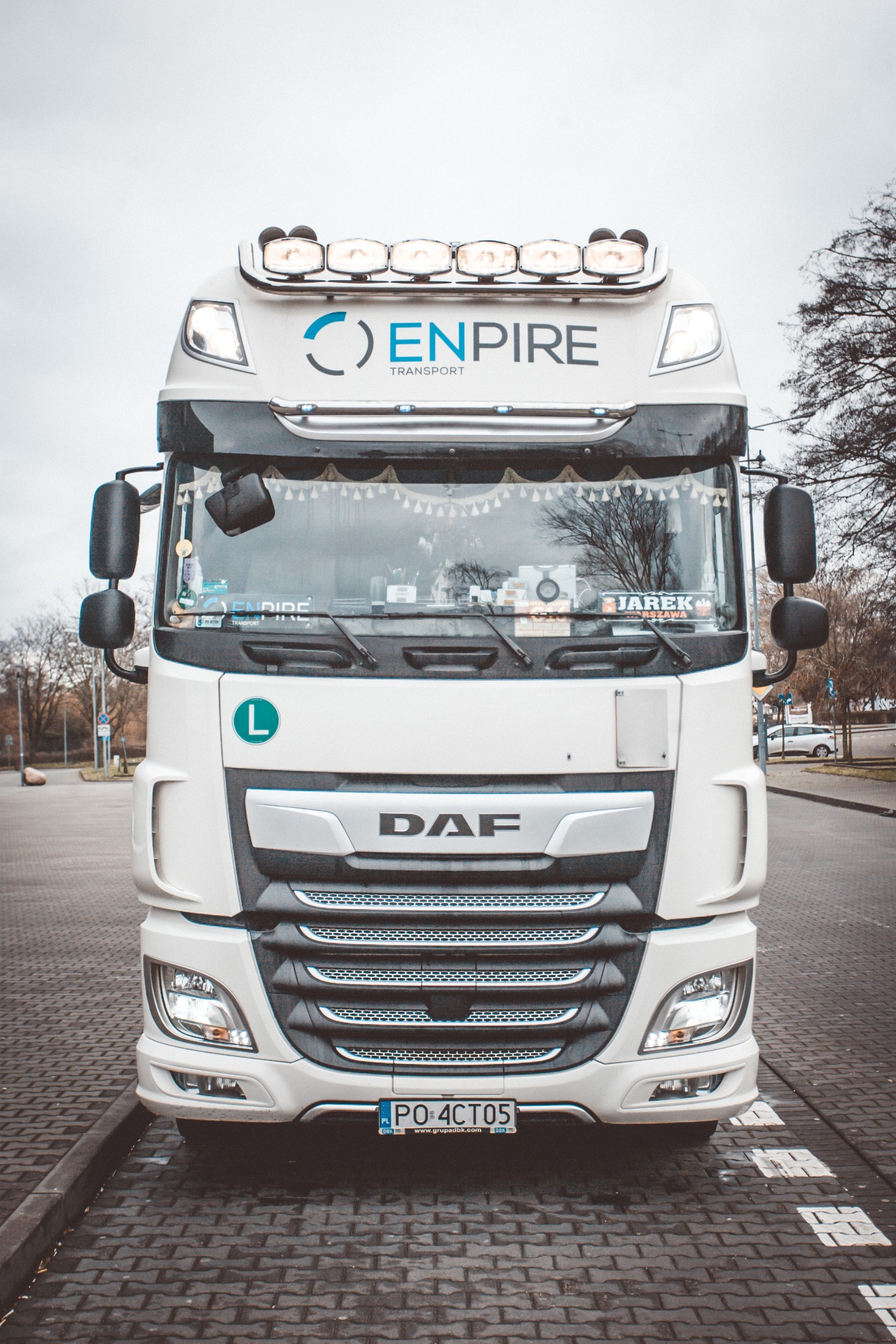 Biała ciężarówka DAF z logo Enpire Transport, tablicą rejestracyjną PO 4CT05 i dodatkowym oświetleniem na dachu, zaparkowana na brukowanej ulicy w pochmurny dzień.